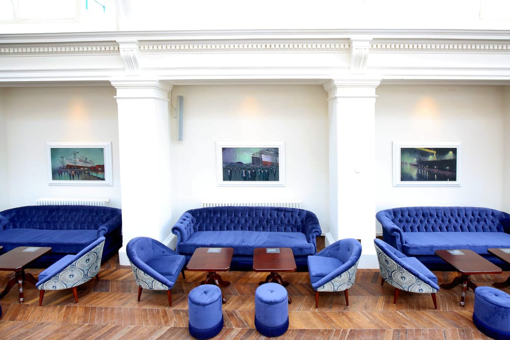 A long lounge lined with blue velvet sofas, low tables and pendant lights above a wooden floor.