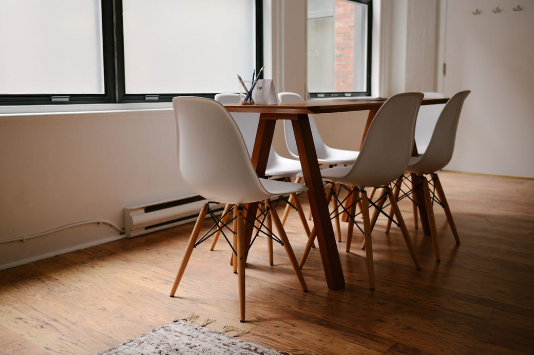 A long wooden table with white moulded chairs standing on a bare wood floor by the window.