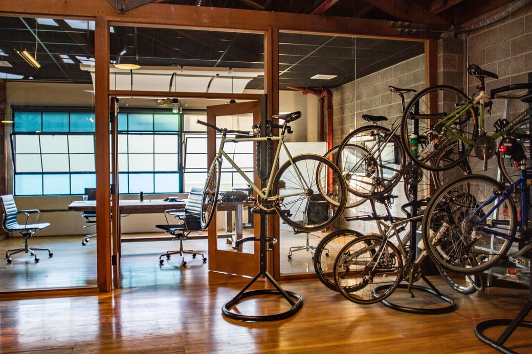 Rows of bicycles racked inside a brick and timber office, with desks visible through glazed partitions.
