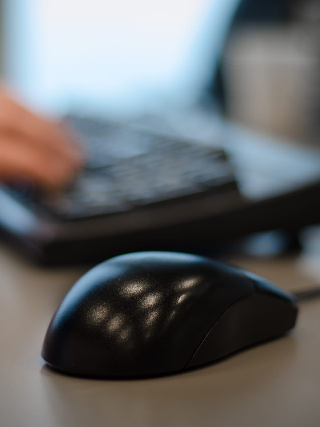 A hand on a computer mouse beside a keyboard and a monitor on a working desk.