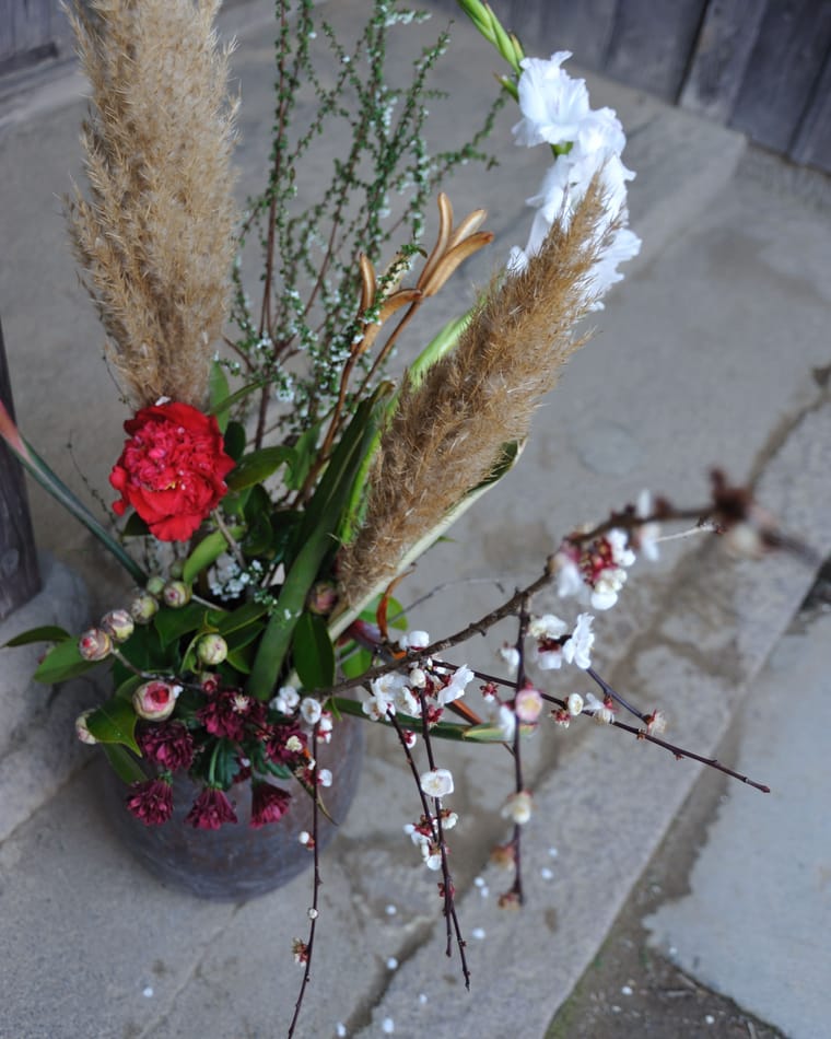 A tall arrangement of pampas grass, a carnation and a blossom branch in a ceramic vase.
