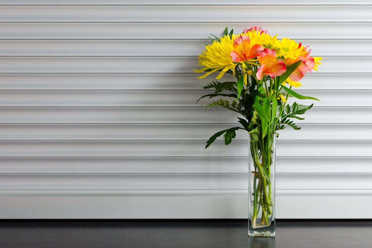 A tall glass vase of yellow and coral alstroemeria standing against a pale panelled wall.