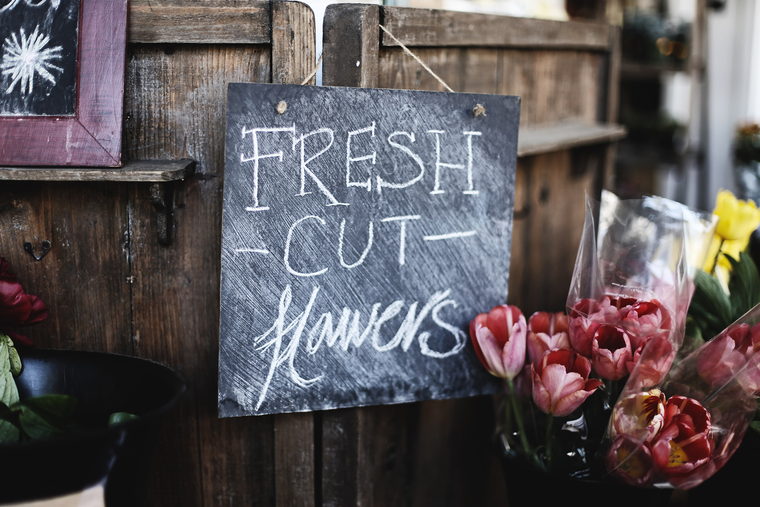 A hand-chalked fresh cut flowers board on a wooden stall, with wrapped tulips in a bucket beside it.