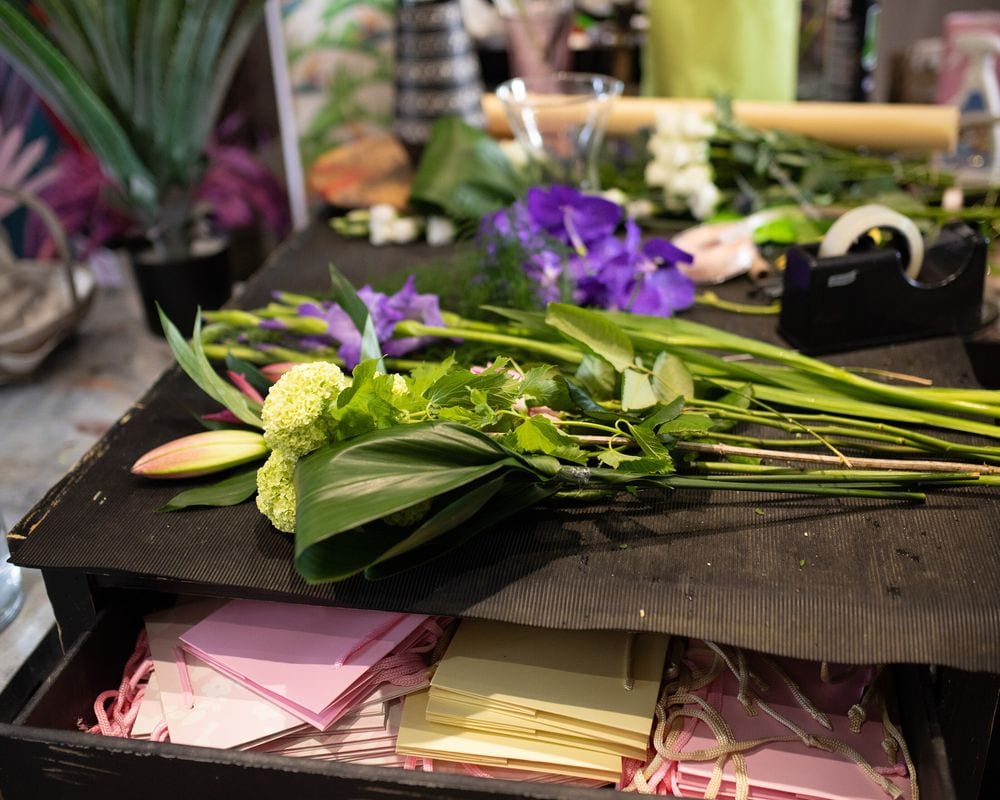 Cut flowers and foliage laid out along a florist's bench, with ribbon and cards in the drawer below.
