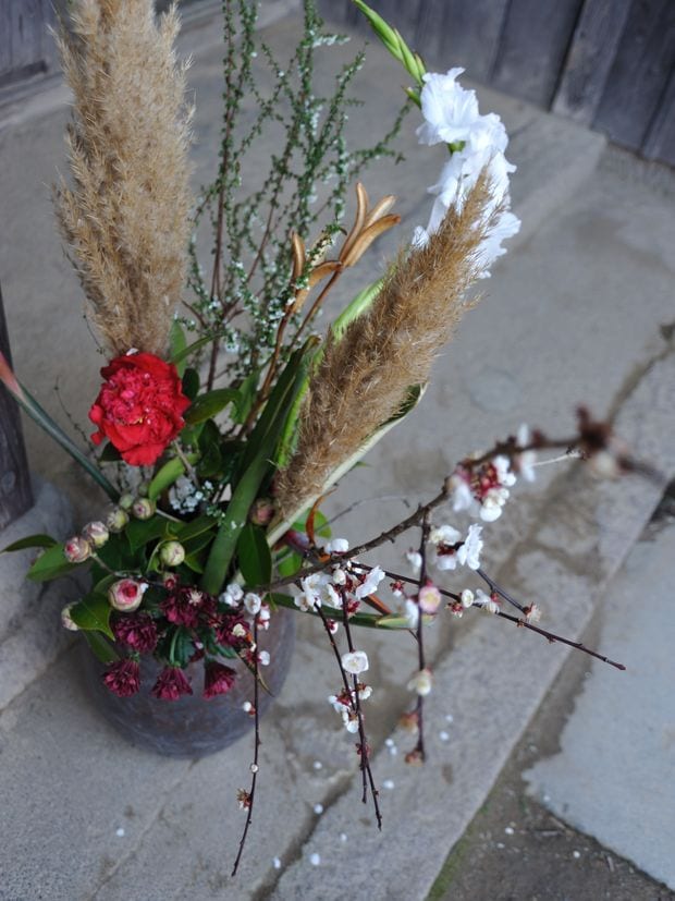 A winter arrangement of pampas grass and a red carnation in a grey pot on a stone step.