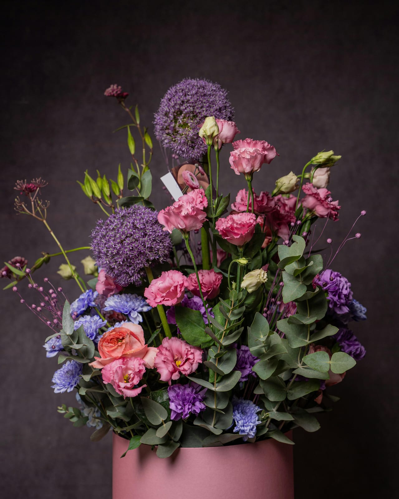 Allium, roses and lisianthus arranged in a pink pot against a dark ground.