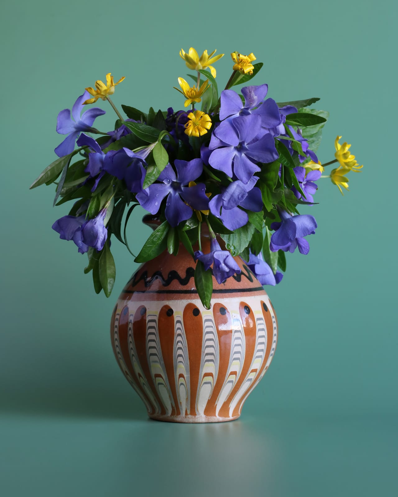 Blue and violet flowers with yellow accents arranged in a striped ceramic jug against a teal ground.