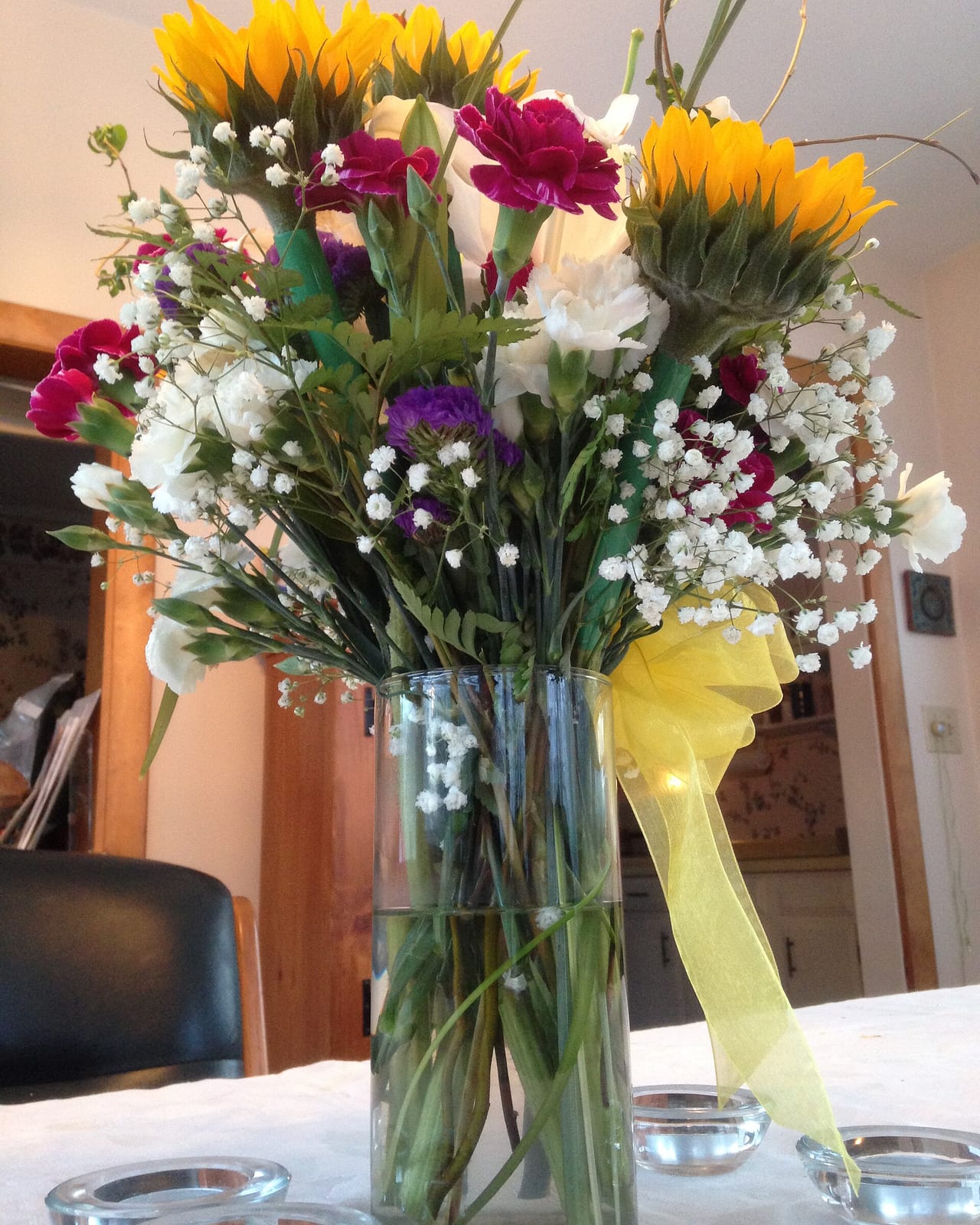 Sunflowers, gerberas, carnations and gypsophila in a tall glass vase finished with a yellow ribbon.