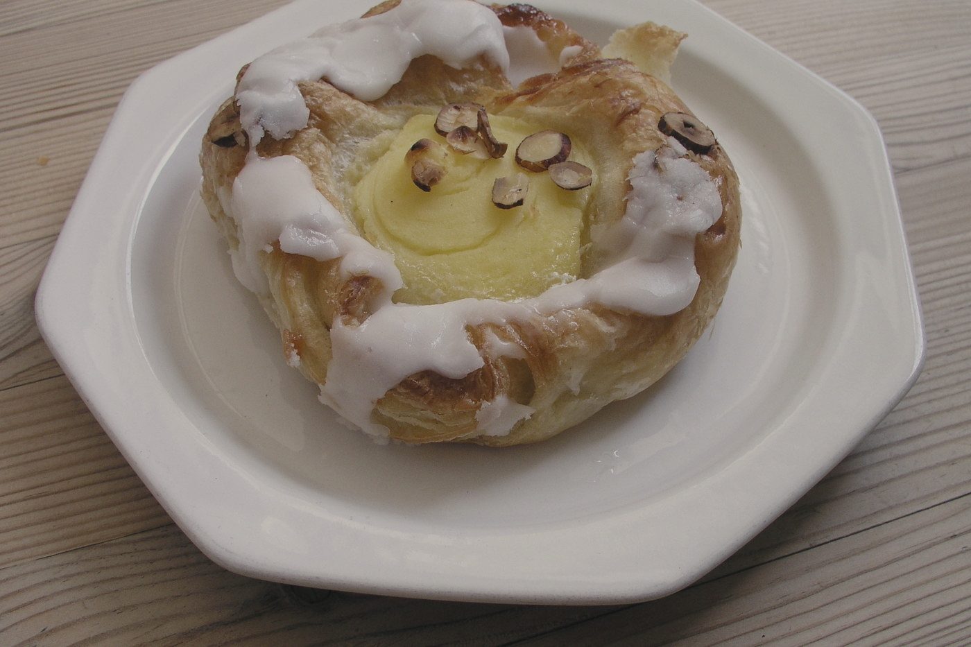 An iced pastry topped with whole nuts on a white octagonal plate on a pale wooden table.