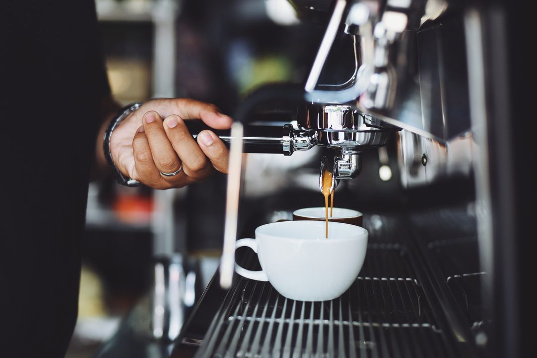 A hand at the group head of an espresso machine as coffee runs into a white cup.