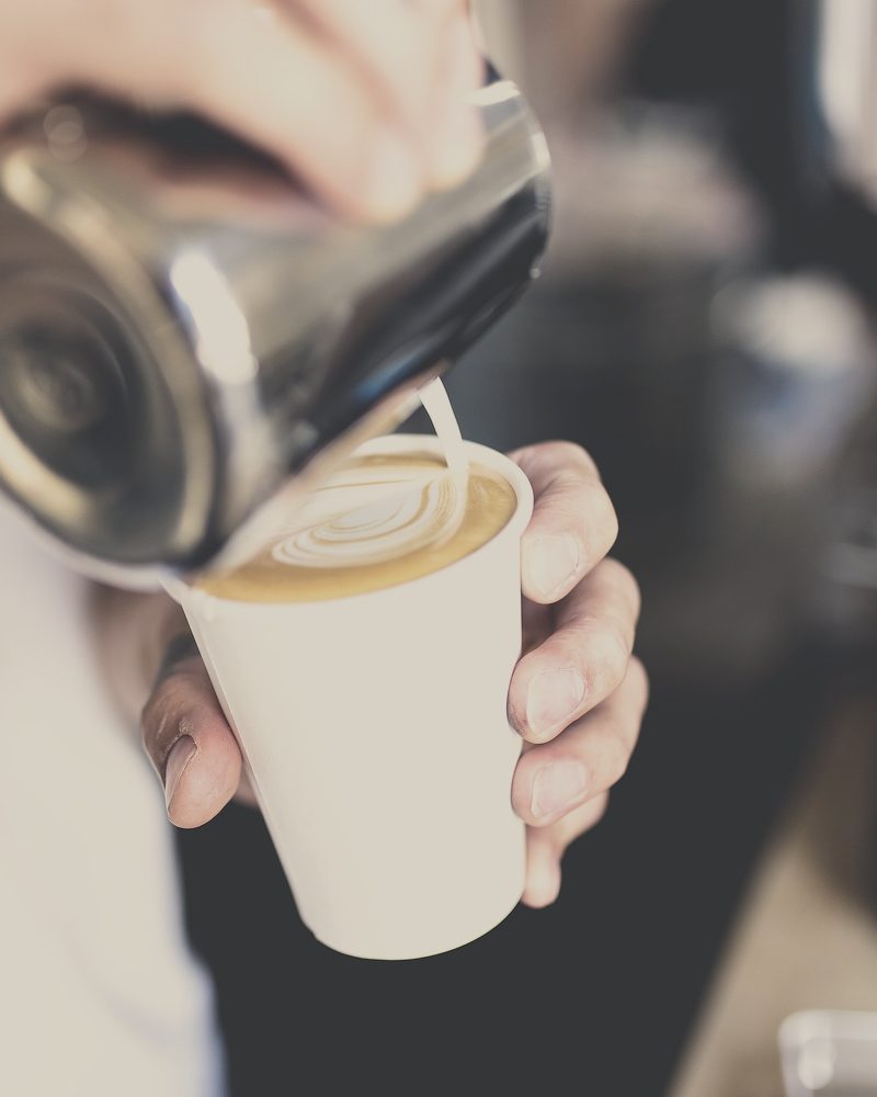 A jug of steamed milk being poured into a cup held in one hand at the counter, photographed from above the shoulder.
