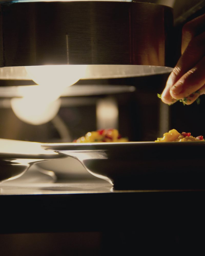 A hand finishing two plates under the low lamp of a kitchen pass, the rest of the room dark behind it.