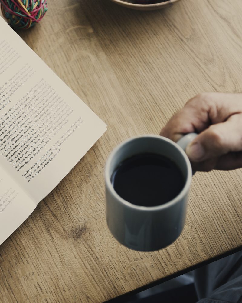 A hand lifting a dark mug of coffee from a wooden table beside an open book, seen from above.