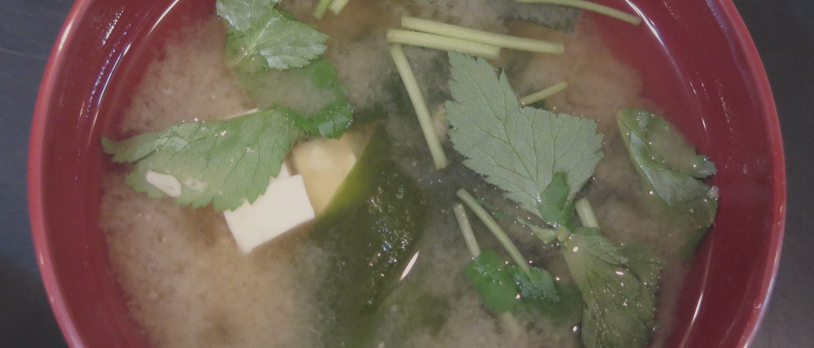 A red lacquer bowl of miso soup with tofu, cucumber and coriander leaves, cropped so the bowl fills the frame.