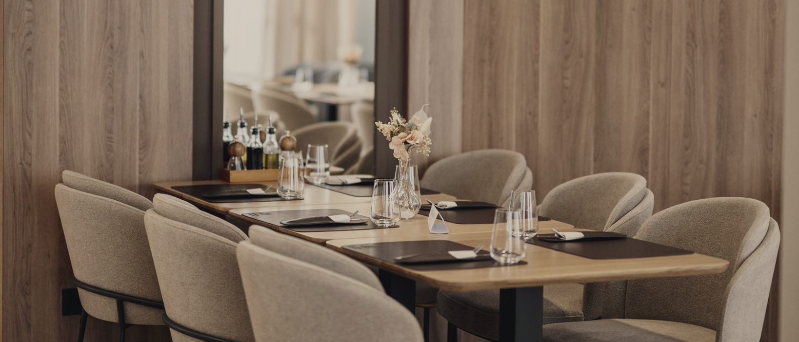A long table laid with mats, glasses and cutlery, ringed by upholstered chairs against a wood-panelled wall.