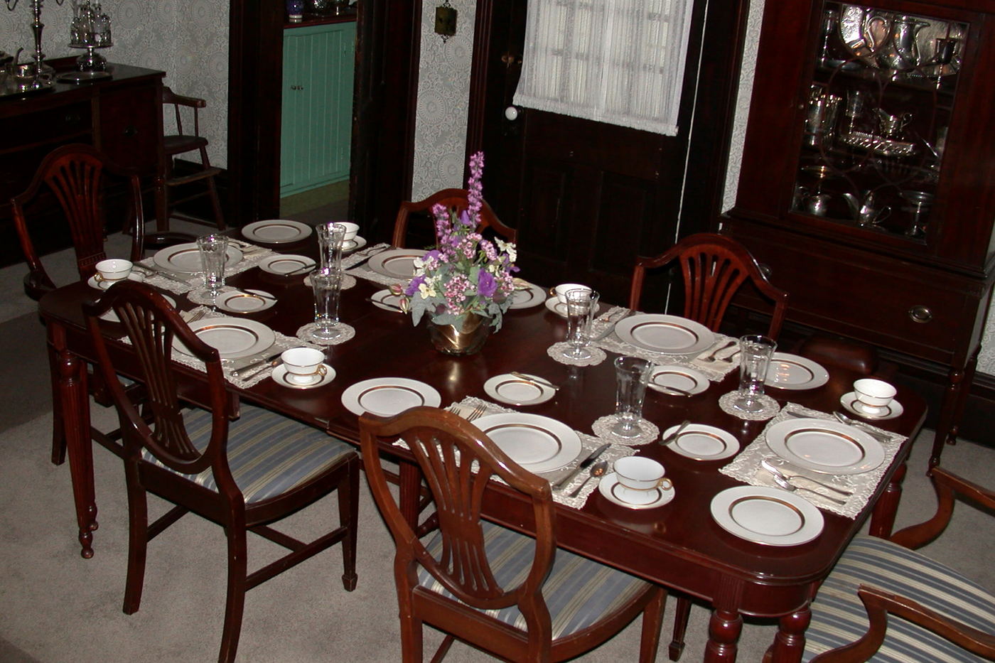 A mahogany dining table laid for six with white and gold china, glassware and a small vase of flowers in the middle.