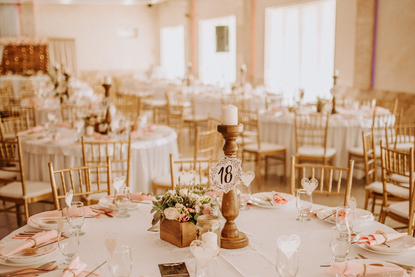 A dressed reception room with round tables, blush napkins, gold chairs and a numbered centrepiece.