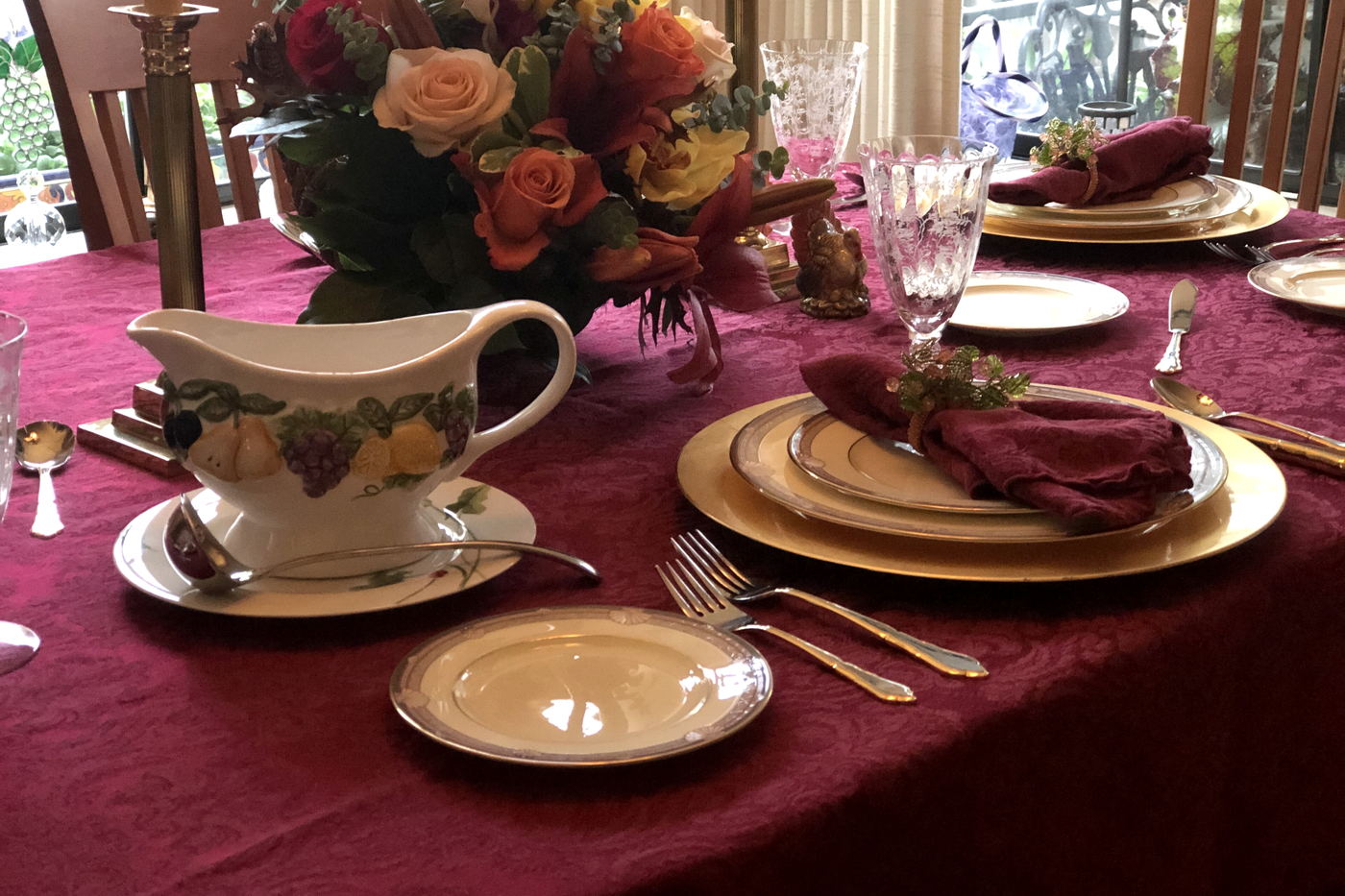 A burgundy cloth laid with gold chargers, folded napkins, cut glass and an autumn arrangement of roses behind.