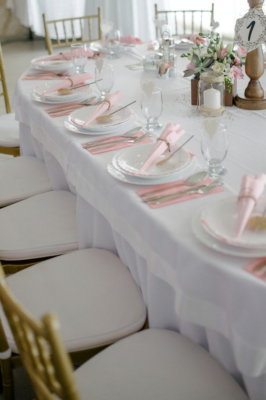 A long banquet table under a white cloth with pink napkins, gold chairs, candle jars and a numbered table card.