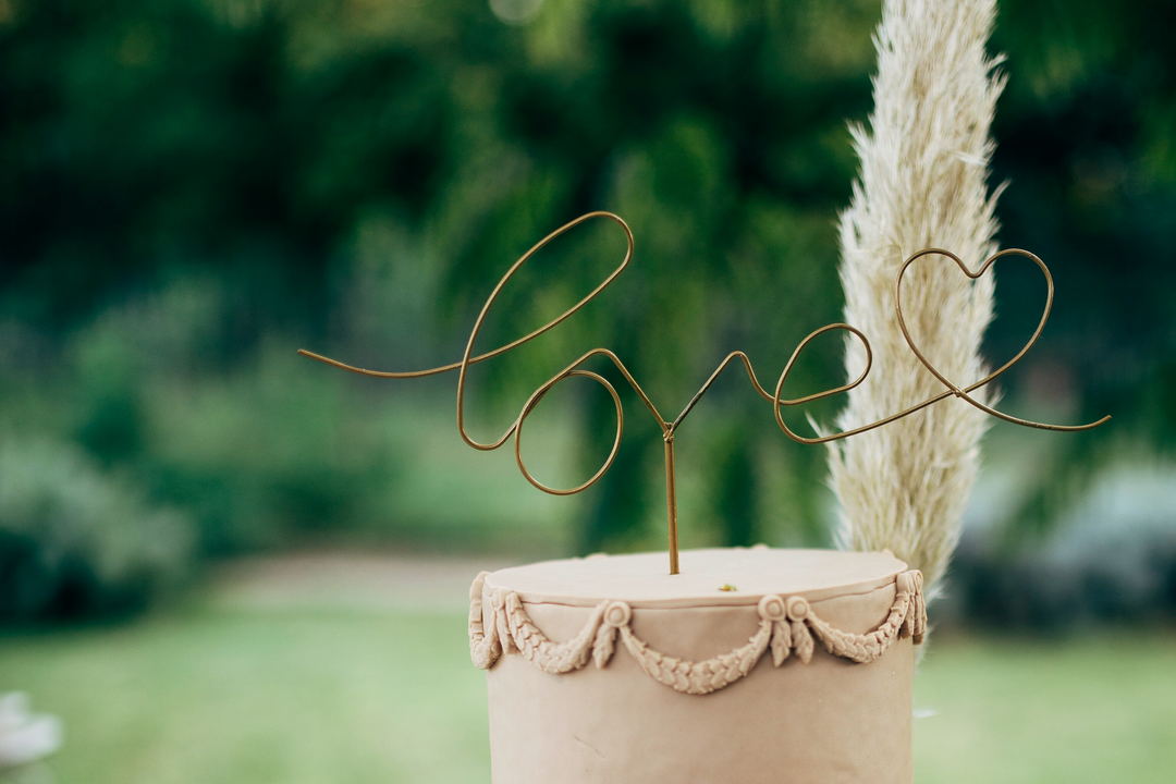 The top tier of a pale cake with rope piping, a wire topper reading love and a plume of pampas grass behind it.