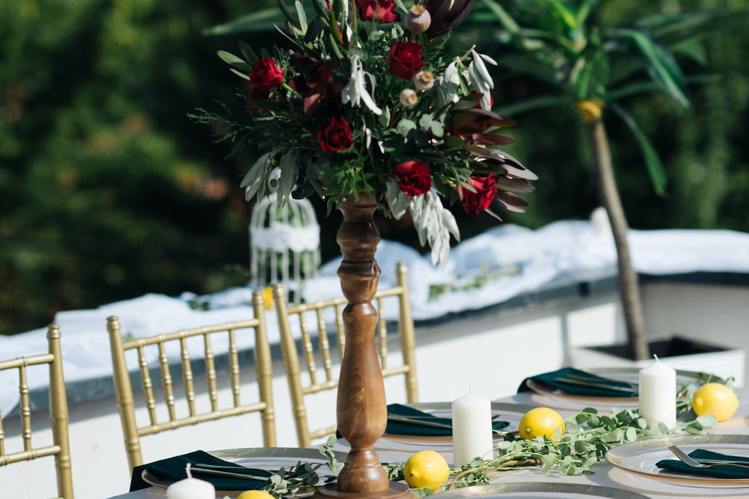A tall arrangement of dark red roses and grey-green foliage on a turned wooden stand above gold chairs, outdoors.
