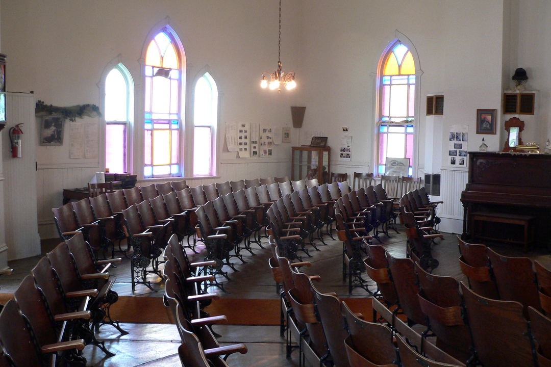 A white-walled hall with pointed stained-glass windows and curved rows of empty wooden seats on cast-iron frames.