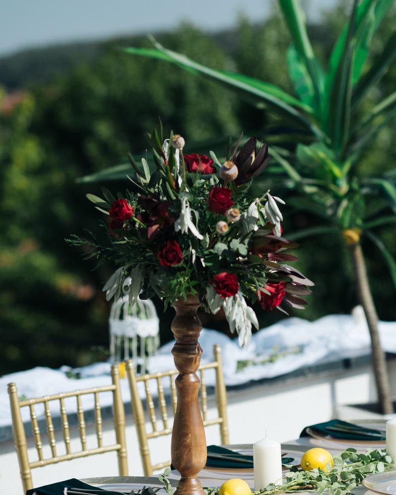 A tall arrangement of dark red roses and grey-green foliage on a turned wooden stand above gold chairs, outdoors.