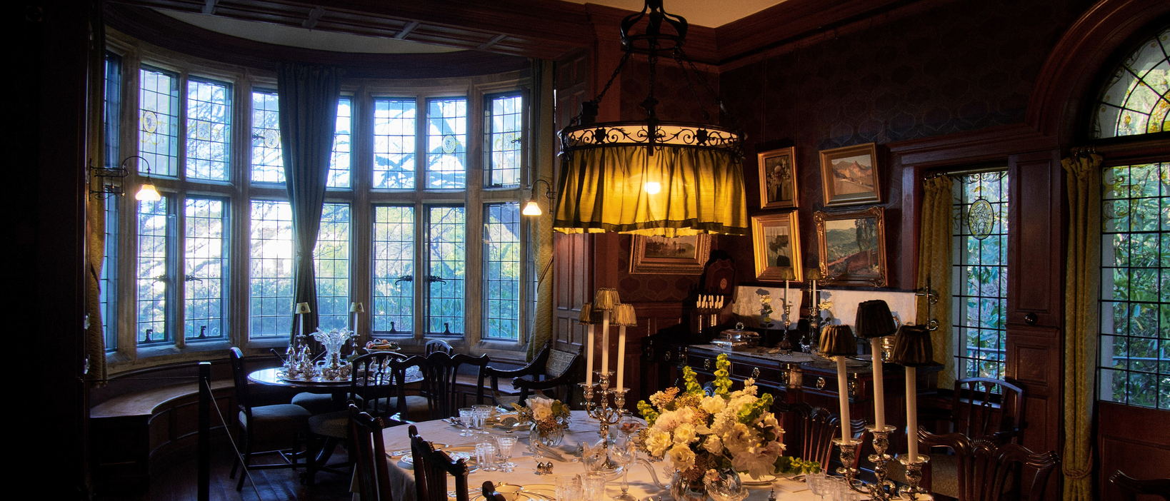 A long table laid in white under a green fringed lamp, against a bay of leaded windows in a panelled room.