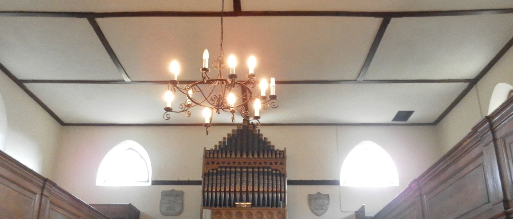 A white beamed ceiling with a wrought-iron chandelier, and a pipe organ on the end wall between two arched windows.