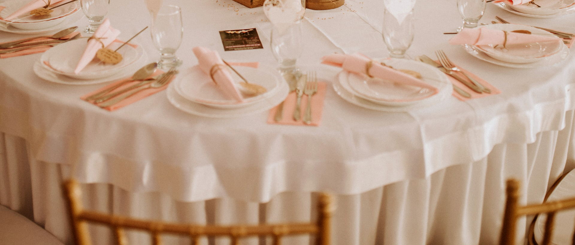 A round table under a white cloth with pink napkins, gold cutlery and gold-rimmed plates, gold chairs in front of it.