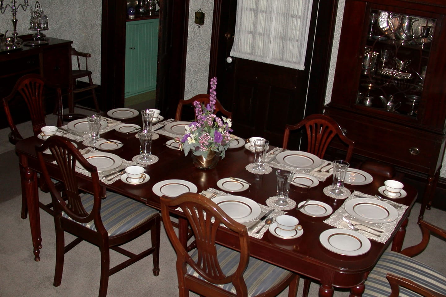 A mahogany dining table laid for eight with white and gold china, cut glass and a small vase of pink flowers.