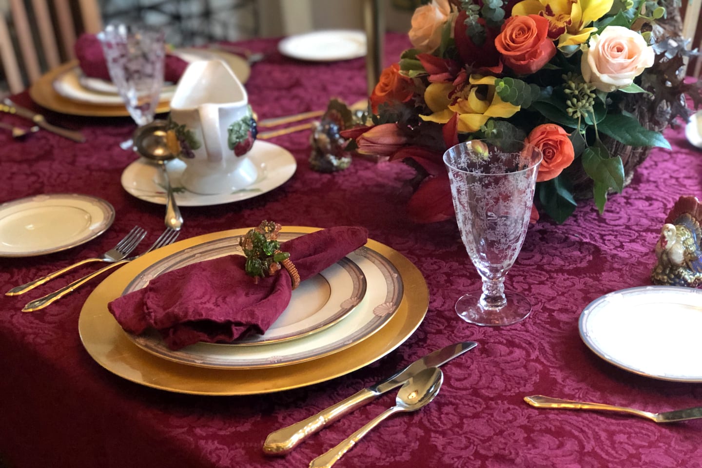 A place setting on a burgundy cloth: a gold charger, a folded napkin in a ring, gold cutlery and an etched crystal goblet.