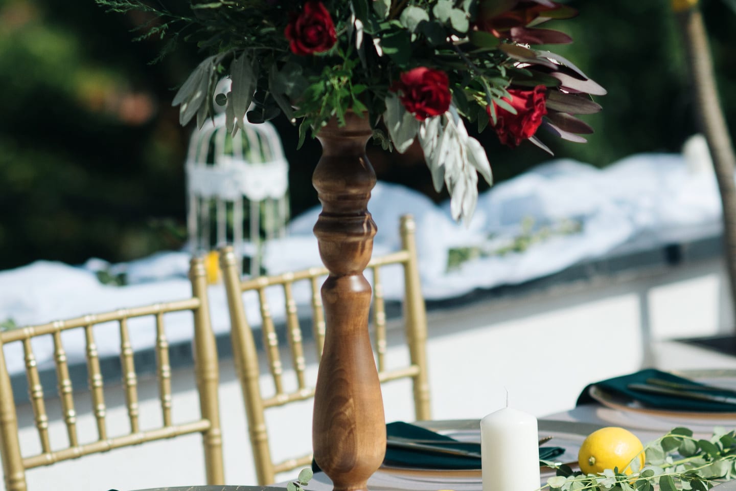A tall arrangement of dark red roses and grey-green foliage on a turned wooden stand above gold chairs.