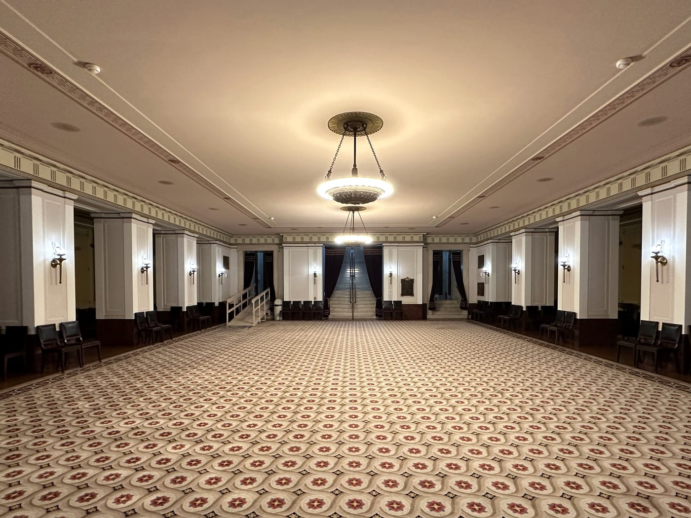 An empty ballroom seen down its length, with a patterned carpet, panelled walls and one ceiling chandelier.
