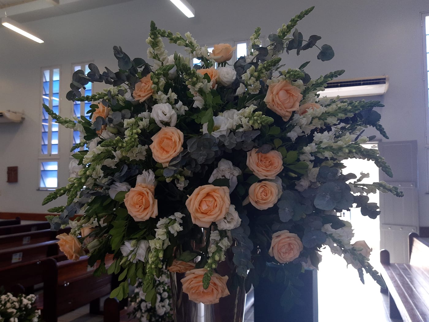 A tall arrangement of peach roses, white stocks and eucalyptus standing beside wooden pews in a chapel.