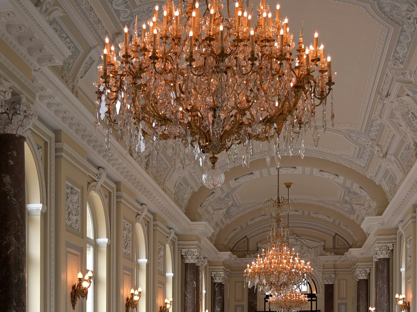 A ballroom ceiling of moulded plaster with two gilt chandeliers hanging in line above dark marble columns.