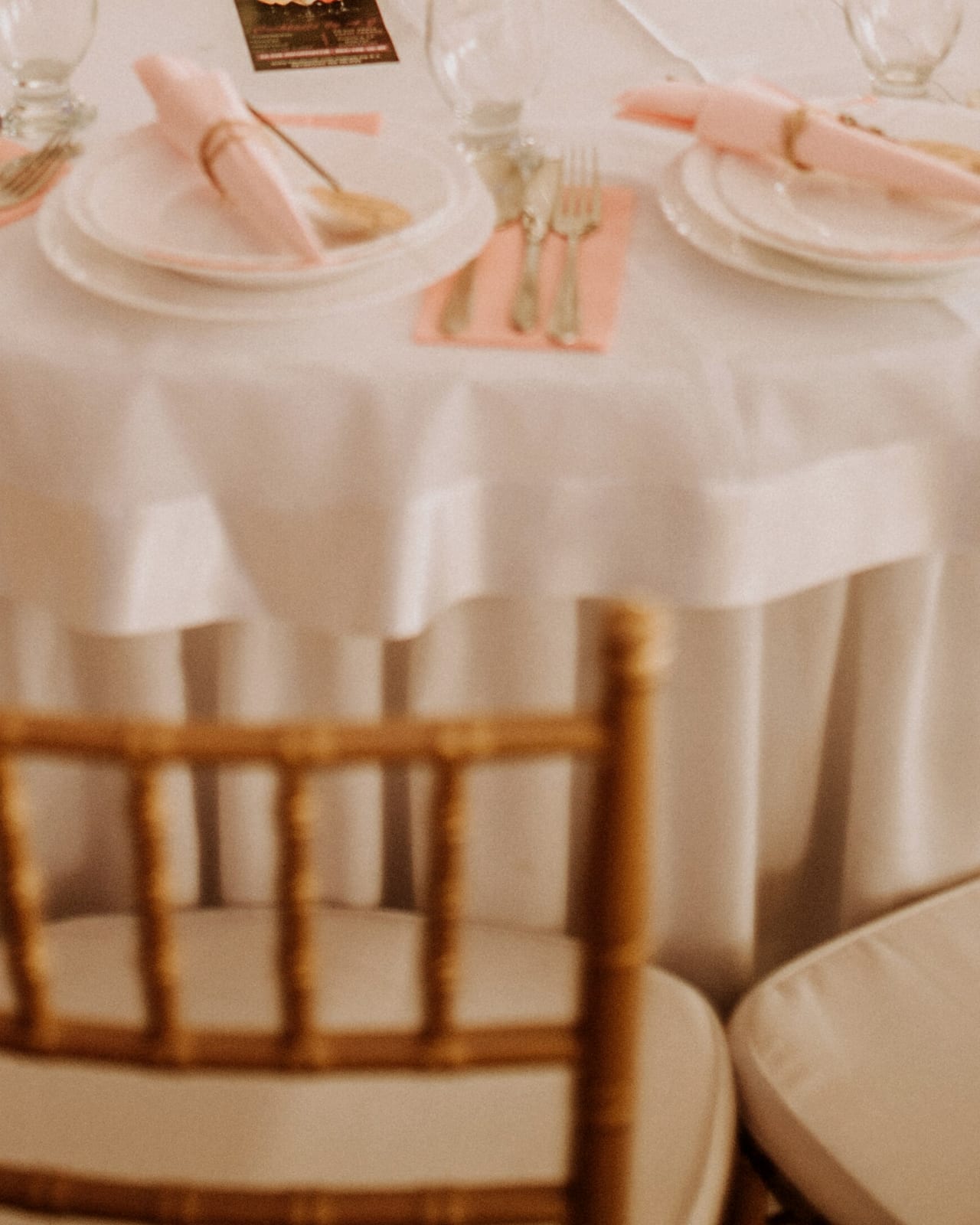 The corner of a round reception table: a white cloth, blush napkins folded on white plates and gold chair backs.