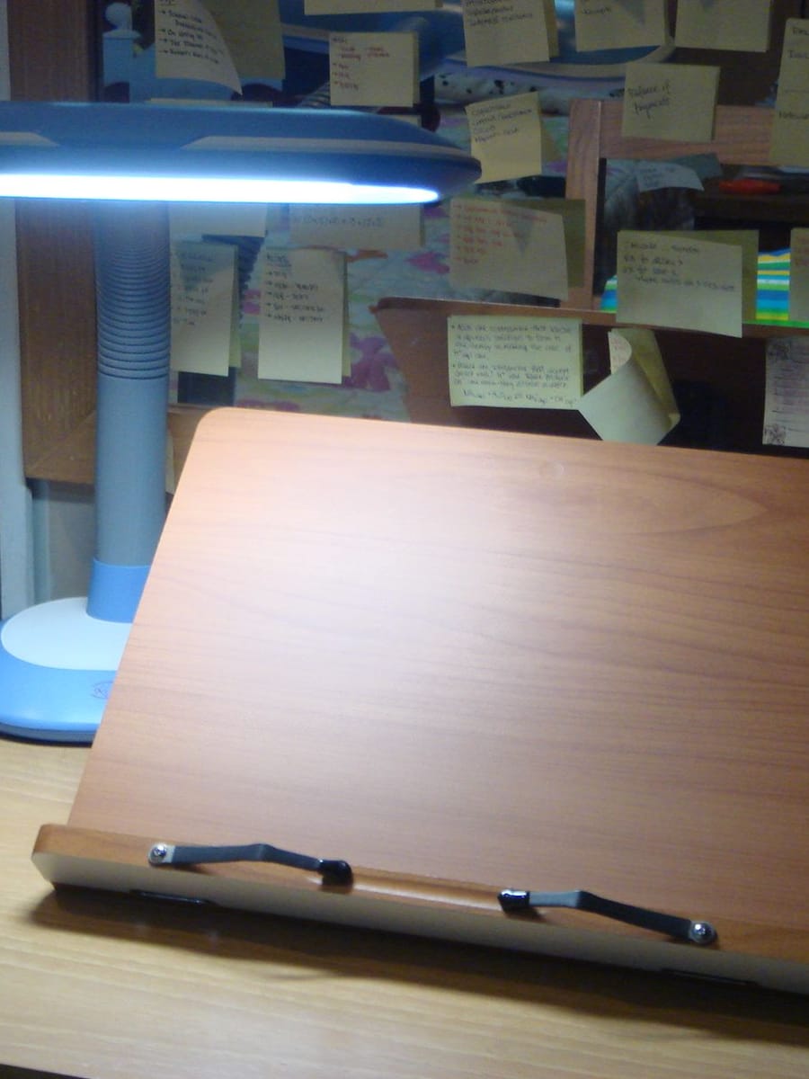 A wooden reading stand on a desk under a blue desk lamp, with handwritten sticky notes covering the wall behind it.