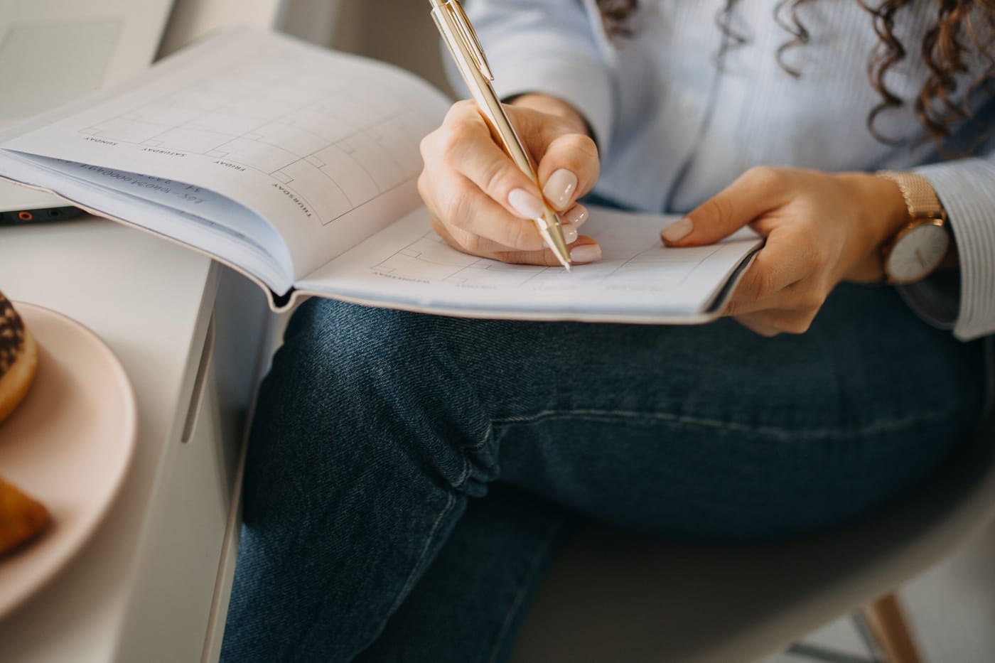 A person writing with a gold pen in a ruled planner resting on their knee, a plate of pastries on the desk beside them.