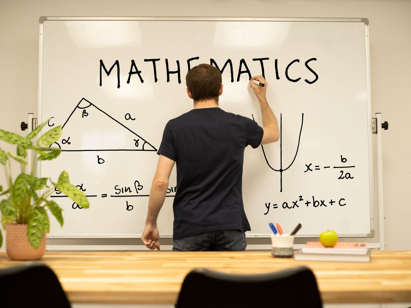 A tutor seen from behind writing MATHEMATICS on a whiteboard beside a labelled triangle, the sine rule and a parabola, with a desk and chairs in the foreground.